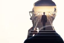 This image features a double exposure effect, blending a close-up profile of a person wearing glasses, deep in thought, with the silhouette of another person walking up a set of stairs towards a bright light, symbolizing contemplation and the journey towards success or enlightenment.