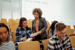 College professor handing back assignment and talking to student about it, with other students sitting nearby