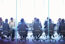 View from behind: about 10 employees sitting at a conference table looking to the right