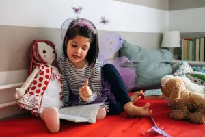 Girl playing alone in her room with her doll, stuffed animal, and book