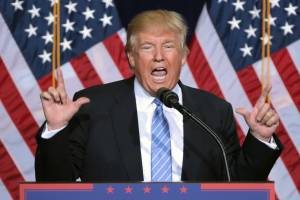 Donald Trump giving a speech with two fingers pointing upward and American flags behind him
