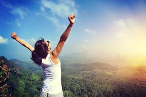 A person stands at the edge of a lush, green valley with arms outstretched towards the sky, celebrating a moment of triumph and freedom. The scene is bathed in sunlight, with a clear blue sky and distant mountains providing a breathtaking backdrop. The person’s white tank top and dark hair are highlighted by the vibrant natural setting.