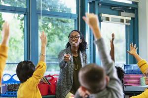 Black teacher calling on a student; lots of students have their hands up
