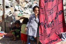 Several children with rubble in the background next to colorful hanging fabric