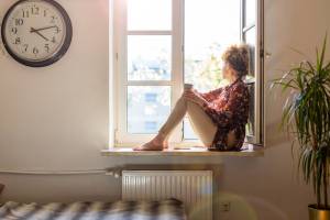 A woman sits on a windowsill, gazing outside with a cup in her hand, bathed in warm sunlight streaming through the open window, creating a serene and contemplative scene.