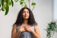 A woman with curly hair is standing with her eyes closed, hands placed on her chest, appearing calm and meditative, surrounded by green plants in a serene setting.