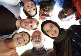 A group of diverse individuals, including men and women of various ages and ethnicities, form a close circle and look down towards the camera, smiling brightly. The photo is taken from a low angle, capturing their joyful expressions and creating a sense of unity and togetherness.