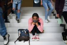 Teen sitting alone on school steps with a mask on, looking depressed