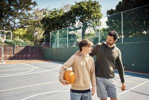 Father and son walking across basketball court; the son is holding a basketball and the father has his arm around the son's shoulders