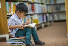 Little boy sitting on a stack of books in the library reading a book