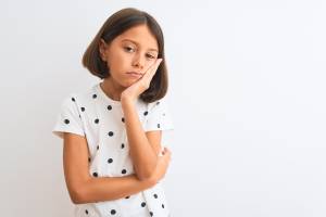 A young girl with a bob haircut, wearing a white shirt with black polka dots, rests her chin on her hand and looks tiredly into the distance, standing against a plain white background.