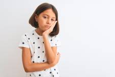A young girl with a bob haircut, wearing a white shirt with black polka dots, rests her chin on her hand and looks tiredly into the distance, standing against a plain white background.