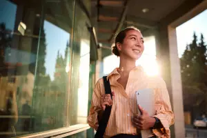 Woman carrying tablet and bag outside office with sun behind her