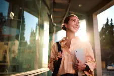 Woman carrying tablet and bag outside office with sun behind her
