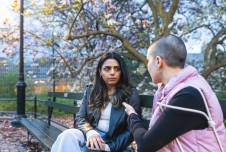 Two people having a serious conversation on a park bench with flowering trees behind them