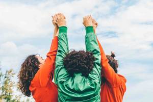 Three people, seen from behind, raise their hands together towards the sky in a gesture of unity and celebration, wearing bright orange and green jackets against a backdrop of a partly cloudy sky.