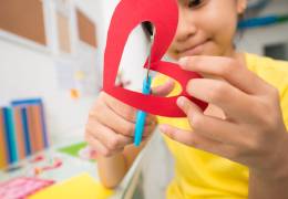 A child is using blue scissors to cut a red paper heart, focusing intently on their craft project at a desk filled with colorful papers and supplies.