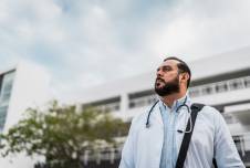 Doctor standing in front of a hospital looking up toward the sky with a tree behind him