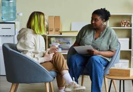 A young person with green-dyed hair, wearing a beige hoodie and brown pants, sits in a modern armchair facing a counselor or therapist. The counselor, an older woman in a green blouse and blue jeans, holds a clipboard and listens attentively. They are in a well-lit office setting with a water dispenser, shelves with books and files, and a tissue box on a small table.