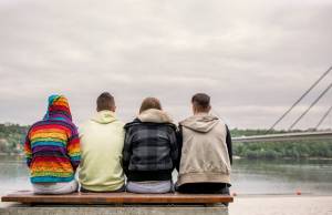 Four people sitting on a bench in front of water and a bridge on overcast day