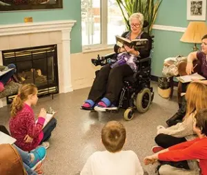 An elder in a wheelchair reading to students
