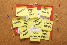 A corkboard is covered with a calendar and numerous yellow sticky notes, each listing different tasks and reminders such as "Dentist," "Pick up kids," "Get milk," "Swim class," "Feed the cat," "Clean the kitchen," "Food shop," "School play," "Dry cleaning," and "Feed the fish." The scene represents a busy and organized schedule with various responsibilities to manage.