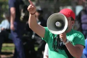 A person wearing a red cap and green shirt is speaking through a megaphone, pointing with their finger, likely addressing a crowd during a public event or protest.