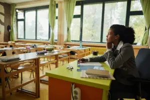 Teacher sitting at her desk looks out over empty classroom with serious expression