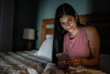 Woman sitting in bed in the dark looking at smartphone and smiling