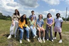 Seven students smiling for a photo in the grass under a cloudy sky