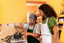 Woman watching her grandmother stir a pot in a bright kitchen