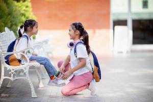 Two girls smiling at each other; one girl is helping the younger girl tie her shoes on a bench