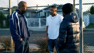 Three men speaking outdoors in front of a fence