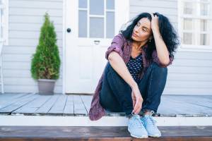 Woman on a porch looking to the side with one hand in her hair and serious expression