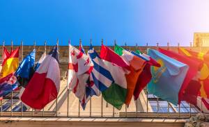 Flags of various countries are displayed on flagpoles in front of a building under a clear blue sky