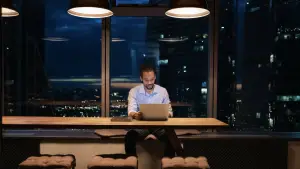 A man working late at night in a modern office with a laptop, surrounded by city lights visible through large windows
