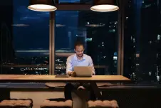 A man working late at night in a modern office with a laptop, surrounded by city lights visible through large windows