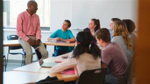 Teacher sitting on a desk talking to circle of students