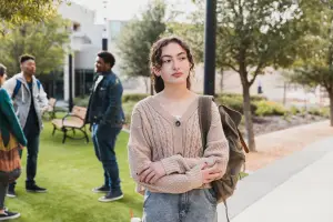 Student standing alone on campus looking hesitant, with other people socializing around her