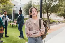 Student standing alone on campus looking hesitant, with other people socializing around her