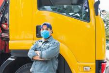 A man wearing a mask stands confidently with arms crossed in front of a yellow truck, likely reflecting resilience during the COVID-19 pandemic