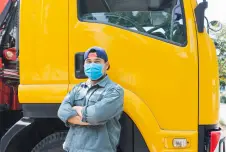 A man wearing a mask stands confidently with arms crossed in front of a yellow truck, likely reflecting resilience during the COVID-19 pandemic
