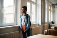 A young boy standing by a window in a classroom, looking outside while holding a notebook and wearing headphones around his neck