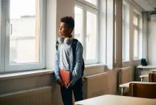 A young boy standing by a window in a classroom, looking outside while holding a notebook and wearing headphones around his neck