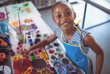 A young girl with a big smile stands at a table covered in art supplies and colorful paintings