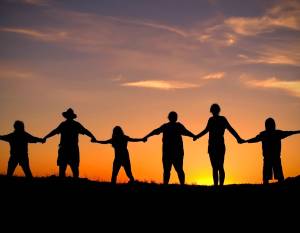 An image showing silhouettes of five people holding hands against a sunset backdrop with warm orange, pink, and purple hues