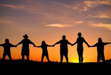 An image showing silhouettes of five people holding hands against a sunset backdrop with warm orange, pink, and purple hues