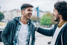 Two men smiling widely at each other outdoors, one with his hand on the other's shoulder