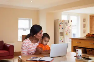 A mother and her young child sit at a table, both looking at a laptop screen
