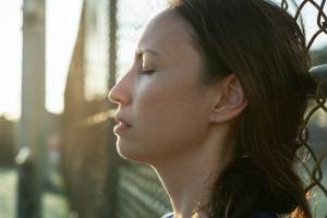 An image showing a woman with closed eyes, appearing to be in a moment of calm or reflection, standing next to a fence with sunlight streaming in